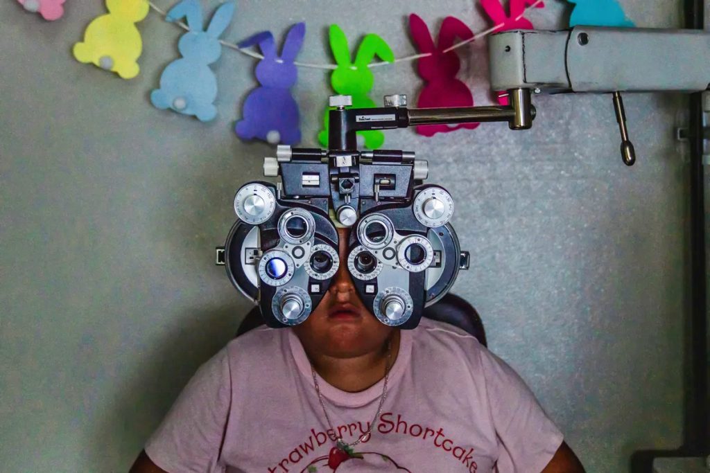 A child, wearing a pink Strawberry shortcake shirt, sits on a chair with their eyes positioned in front of a metal Phoropter during an eye exam.
