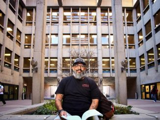 A bearded person wearing a black “Chico State” T-shirt and cap sits on a low concrete planter holding papers, posed in a courtyard framed by tall concrete columns and rows of windows, with a library entrance visible behind them and a few people walking through the background.