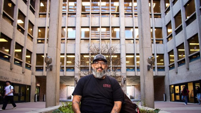 A bearded person wearing a black “Chico State” T-shirt and cap sits on a low concrete planter holding papers, posed in a courtyard framed by tall concrete columns and rows of windows, with a library entrance visible behind them and a few people walking through the background.